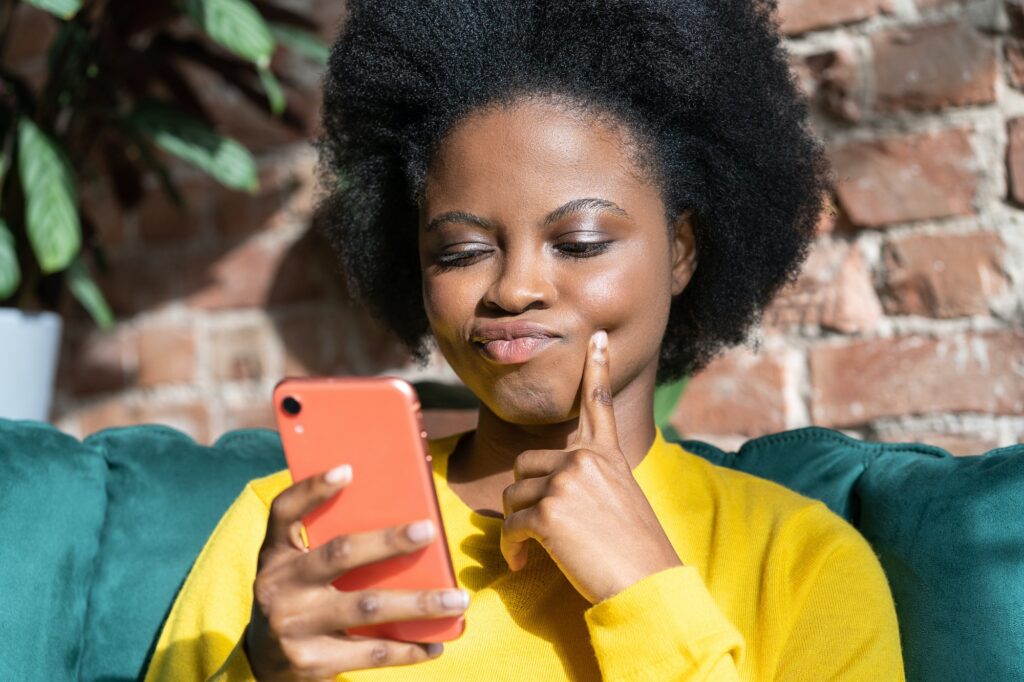 Black woman ponders on how to answer question, using mobile phone, tries to made up good message