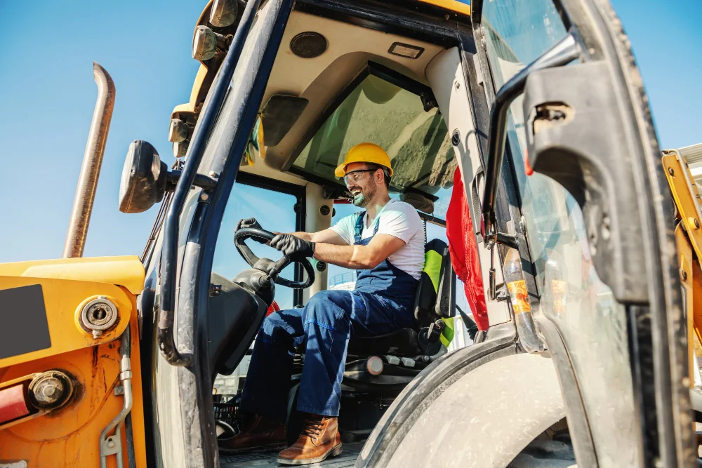 Smiling handsome caucasian worker in overall and with helmet on head driving excavator